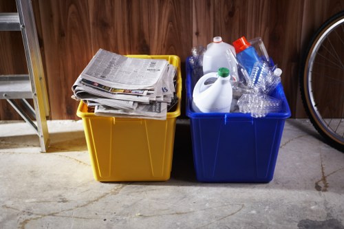 In a residential garage or utility room, two large plastic recycling bins are positioned on a concrete floor against a wooden paneled wall. The yellow bin on the left is filled with neatly stacked and slightly crumpled newspapers and magazines, with some pages sticking out over the edge. The blue bin on the right contains various recyclable plastic bottles of different sizes and shapes, some featuring caps, and a few clear plastic containers, all loosely arranged. The bins are situated next to each other, with the yellow one appearing slightly closer in the foreground and the blue bin behind it. To the right of the bins, part of a bicycle wheel is visible, indicating a multi-purpose storage space. The lighting is soft and natural, highlighting the textures of the plastic, paper, and concrete, and subtly illustrating the setting as part of a typical household environment, in an area where rubbish and recyclables are stored before disposal or collection by local waste management services in Ealing or surrounding areas.