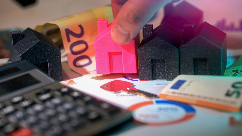 A close-up view of a hand holding a small pink model house made of plastic, positioned among three larger black model houses with a textured finish, resembling rooftop structures, on a flat surface. T