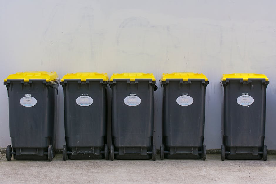 Three large black plastic trash bags filled with household waste are placed on a pavement next to a curb. The bags appear to be full and are slightly crumpled, with some writing visible on them, indic