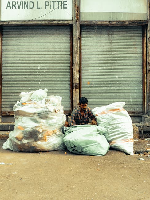 A group of five individuals working in a large open landfill site surrounded by high rocky walls, dumping and collecting mixed waste materials onto the ground. The waste consists of plastic, paper, fa